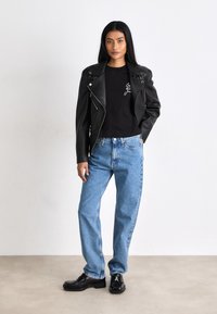 Chaqueta de motorista de cuero negro con cremalleras plateadas, camiseta negra con gráfico blanco, jeans rectos de mezclilla azul claro y zapatos negros.