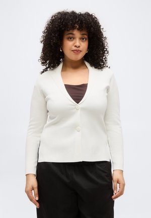 Woman with curly hair wearing gold hoop earrings, a white buttoned cardigan over a brown top, and black pants standing against a plain background.