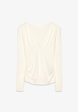 Kreemist pikakäiselise särgiga, millel on sügav V-kaelus, kohandatud detailid bodice'il ja varrukatel ning pehme ja elastne kangas.