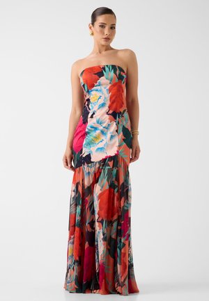 Femme portant une robe longue sans bretelles à fleurs larges et colorées en rouge, bleu et vert, debout devant un fond uni.