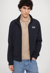 Chaqueta de marina con cuello alzado, cierre de cremallera, puños elásticos y un logo blanco. Lleva puesta sobre una polo beige, combinada con jeans azules.