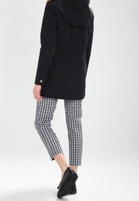 Svart huva jacka med guldknappar, i kombination med svart-vit rutiga byxor och svarta skor. Enkel, strukturerad design.