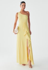Yellow one-shoulder dress with textured fabric, ruffled side detail, and high slit. Paired with a small white handbag and nude heels.