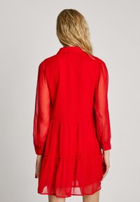 Robe rouge avec un design en couches, manches longues en dentelle et col. Un tissu léger crée une silhouette fluide. Manches à boutons.