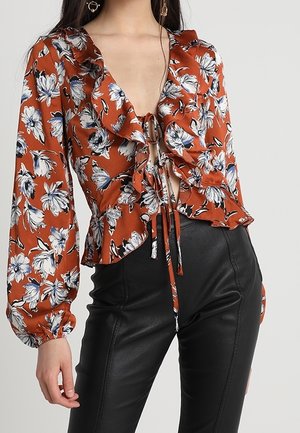 Roest-oranje blouse met bloemenprint, geruchte kraag, lange pofmouwen, detail met strik aan de voorkant, gecombineerd met zwarte leren broek met hoge taille.