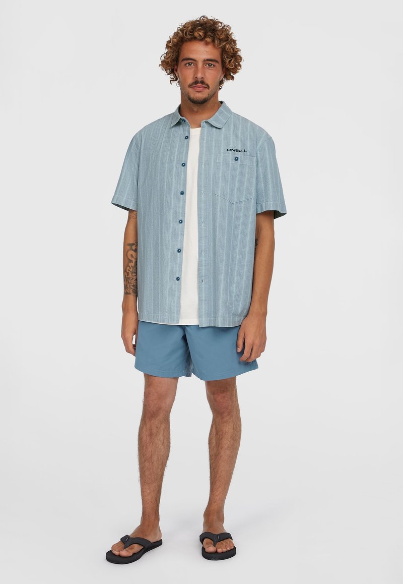Kortærmet, knap-op skjorte med et vertikalt blåt stribe mønster, parret med lyseblå shorts og sorte flip-flops. Har en brystlomme.