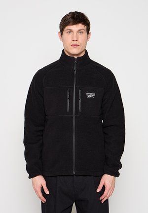 Uomo che indossa una giacca in pile nera Reebok con zip intera e due tasche verticali sul petto, in piedi davanti a uno sfondo chiaro e uniforme.