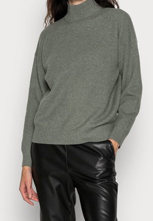 Maglione verde a maglia con collo alto, spalle scese e polsi e orlo a coste, abbinato a pantaloni neri in ecopelle.