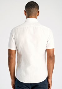 Man met zijn rug naar de kijker, gekleed in een wit shirt met korte mouwen en donkere broek, tegen een effen lichte achtergrond.