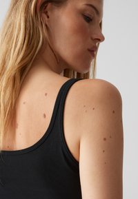 Top nero senza maniche con spalline sottili, caratterizzato da una trama liscia. La pelle della modella mostra vari piccoli segni sulla parte superiore della schiena e della spalla.