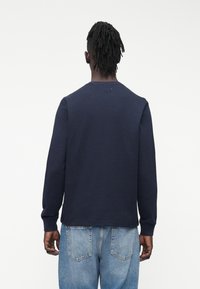 Sinine lahtiste varrukatega triiksärk soonilise tekstuuriga, ümar kaelus ja kitsenevad varrukasäärised, kombineeritud heleroheliste teksapükstega. Tagantvaade.