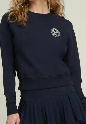 Kvinde iført en navyblå langærmet sweatshirt med et lille cirkulært hvidt logo på venstre bryst og et matchende plisseret nederdel.