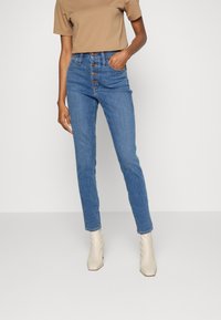 Jeans de mezclilla azul de cintura alta con cierre de botones y dos bolsillos laterales. Combinados con una blusa corta de manga corta color beige y botas blancas hasta el tobillo.