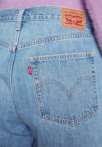 Jeans de mezclilla azul claro con una etiqueta de cuero de marca, que presentan un distintivo bordado de doble arco en el bolsillo trasero y un detalle de etiqueta roja.
