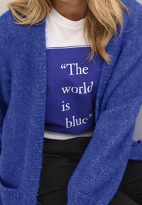 Cardigan de punto azul superpuesto a una camiseta blanca con un estampado azul que presenta el texto: "El mundo es azul". Tela negra en la cintura.