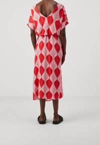 Robe à motifs de feuilles rouges et blanches avec un dos en V, une coupe décontractée, des manches courtes et une longueur jusqu'aux genoux. Portée avec des sandales noires.