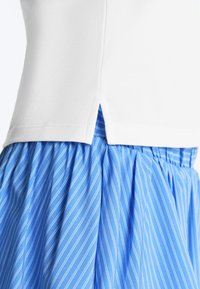 Haut blanc avec fente latérale porté sur une jupe bleue à rayures blanches diagonales et taille froncée.