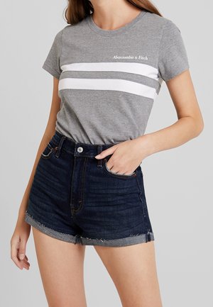 Woman wearing grey Abercrombie & Fitch t-shirt with white stripes and dark blue rolled-up denim shorts, hand in pocket.
