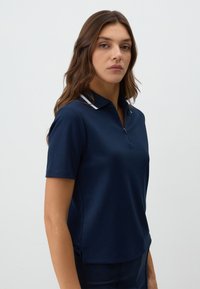Marineblauwe polo met korte mouwen, ritssluiting en witte kraagaccenten. Gemaakt van een gladde, lichte stof met een ontspannen pasvorm.