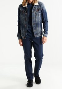 Jeansjacka med fårskinnskrage och framfickor, kombinerad med en mörkblå långärmad skjorta och marinblå byxor, komplett med svarta sneakers.