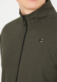 Tummanvihreä vetoketjullinen huppari, jossa on korkea kaulus, musta vetoketju ja pieni logotyyppinen yksityiskohta. Valmistettu pehmeästä kankaasta, jossa on sileä pinta.