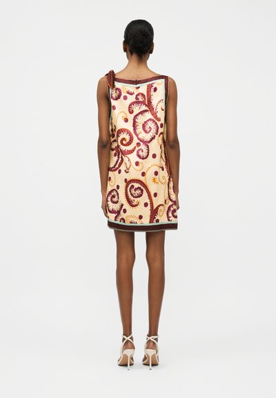 Γυναίκα που στέκεται με την πλάτη προς τον θεατή, φορώντας αμάνικο μπεζ φόρεμα με μπορντό και πορτοκαλί κυματιστό μοτίβο και λευκά σανδάλια με ψηλά τακούνια.