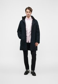 Fiatal férfi hosszú fekete kabátot visel, alatta világos rózsaszín gombos ing van, fekete nadrág és fekete francia koptatott cipő fehér zoknival.