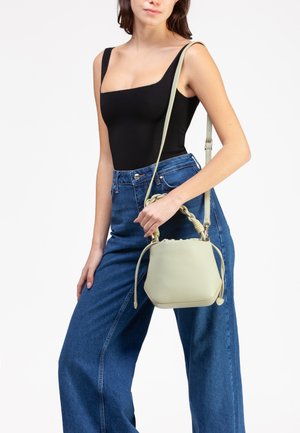 Donna che indossa un top nero senza maniche e jeans blu a gamba larga con vita alta, tiene in mano una borsa a secchiello beige con manico intrecciato e tracolla.