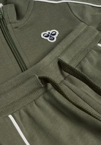 Olivengrøn hoodie med lynlås og hvide detaljer. Har et tekstureret stof og et broderet bi-logo på brystet. Strækbart taljebånd.