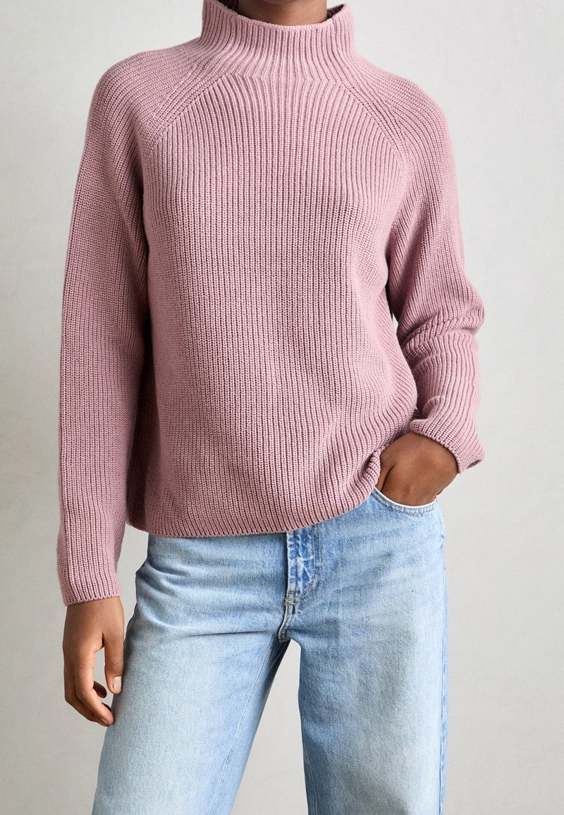 Pink ribbed knit sweater with a high collar and relaxed fit, paired with light blue denim jeans. Sweater features long sleeves and textured fabric.