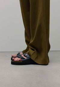 Zwarte sandalen met twee gespen en een kurken voetbed, gedragen met losse, bruine broeken, waarbij de tenen zichtbaar zijn. Een gladde textuur en een platte rubberen zool.