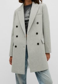 Lichtgrijze dubbelgeknoopte wollen jas met zwarte knopen, gedragen over een zwart shirt en lichtblauwe jeans.