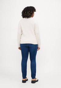 Lys beige teksturert genser med ribbet kant, sammen med mørkeblå slim-fit jeans. Brukt med svarte platåsandaler. Utsikt bakfra.