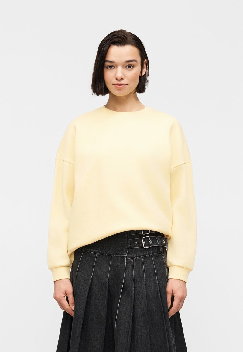 Jeune femme aux cheveux courts et foncés portant un sweat-shirt jaune pâle et une jupe en denim foncé plissée avec un détail à double boucle.