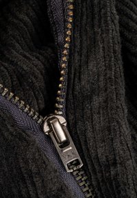 Schwarzer Cordstoff mit einer Nahaufnahme eines metallischen Reißverschlusses mit YKK-Zug; die Textur ist gerippt und weich.