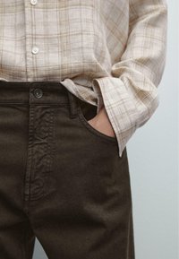 Světle kostkovaná košile s hnědými a krémovými barvami, spojená s tmavě hnědými kalhotami s přední kapsou a detailním kovovým knoflíkem.