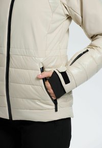 Polstret beige jakke med en blank finish, der har lynlåslommer, sorte accenter og manchetter samt et slankt, moderne design.