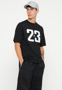 Melns T-krekls ar lielu baltu "23" un logo grafiku; melnas šortus ar sarkanu akcentu; gaiši pelēka cepure. Ikdienišķais sportiskais stils.