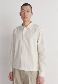 Camicia chiara a maniche lunghe con righe verticali, apertura frontale con bottoni e una texture morbida. Presenta un colletto classico e una vestibilità rilassata.