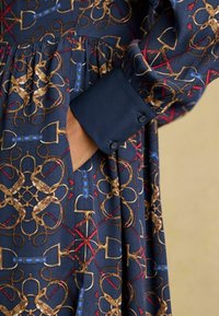 Tumes sinise riide kangas sisaldab kuldset ja punast ratsutamismustrit, lähedalt vaadates on näha nuppega mansetti, mis on valmistatud ühtlasest tumesinisest kangast, rõhutades tekstuuri ja disaini detaile.