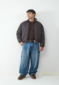 Puffermantel in Dunkelbraun, strukturierter Strickpullover, lockere blaue Jeans, braune Schuhe mit weißer Naht, kontrastierender Gürtel.