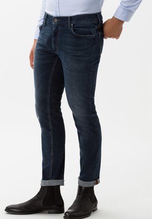 Jeans de mezclilla oscura con corte ajustado, con cierre de botón, cinco bolsillos y un detalle de puño enrollado. Combinados con botas de tobillo negras.