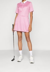 Krótka, różowa sukienka golfowa w paski z krótkim rękawem i białym kołnierzykiem; zestawiona z białymi butami i kijem golfowym trzymanym w lewej ręce.
