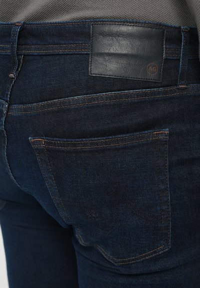 Close-up of dark blue denim jeans back pocket and black leather brand patch on waist, stitched with orange thread.