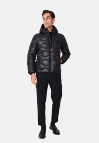 Giacca a vento nera con design trapuntato, cappuccio e chiusura con zipper. Indossata su pantaloni neri e scarpe nere, con una tasca laterale.