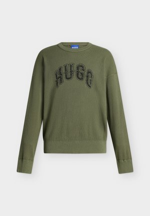 Jeune homme debout avec une main dans la poche, portant un pull vert, un jean bleu, des chaussures noires et portant un sac à bandoulière marron.
