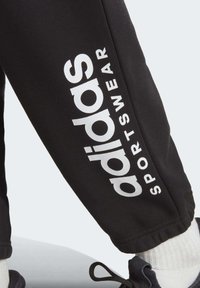 Pantaloni sportivi neri con un prominente logo "adidas" bianco e la scritta "SPORTSWEAR" lungo il lato, realizzati in un tessuto morbido e texture.
