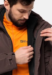 Giacca impermeabile marrone che si apre per rivelare uno strato interno in pile arancione con cerniera. Presenta un logo sul pile e cinturini regolabili sui polsini.