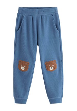 Kind trägt blauen Pullover und Hose mit Teddybär-Gesichtern, richtet einen karierten Hut, steht vor schlicht hellem Hintergrund.