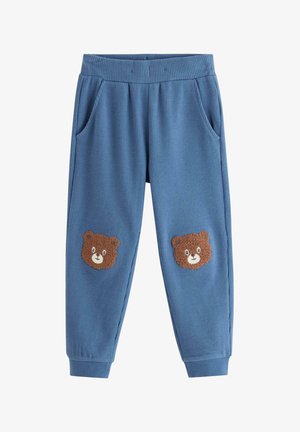 Barn iført blå sweatshirt og bukser med bamseansigter, justerer en ternet hat, står på en ensfarvet lys baggrund.